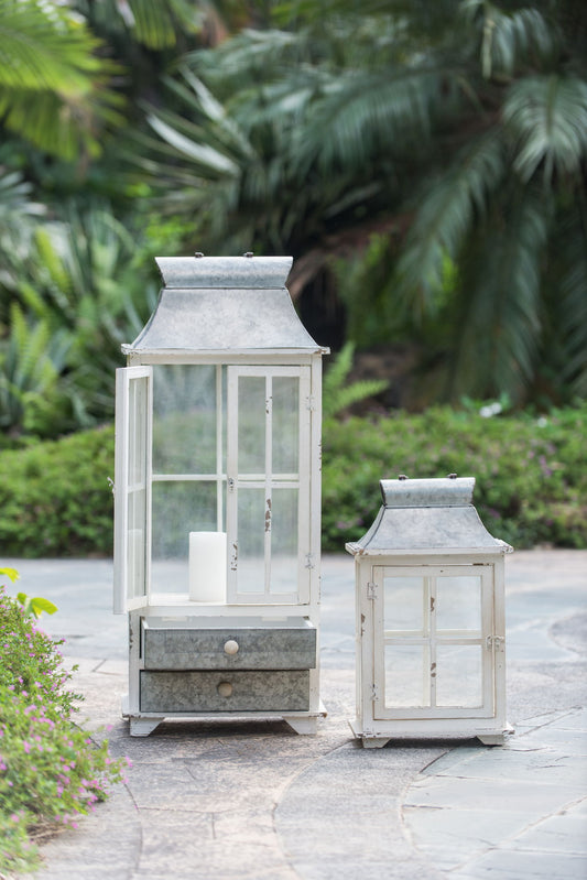 The Wooden Candle Lantern Decorative, Hurricane Lantern Holder Decor in ivory stands on a stone path amid lush greenery and tropical plants, featuring one large lantern with a white candle and drawer, and one smaller, both with glass panels.