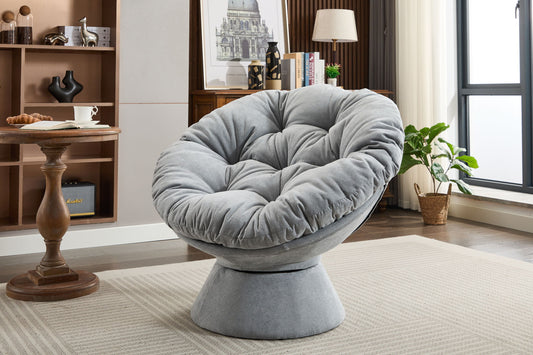 A modern living room with an Oversized Swivel Accent Chair, 360 Swivel Barrel Papasan Chair on a beige rug, complemented by a bookshelf, wooden side table, potted plants, and a large window in the background.