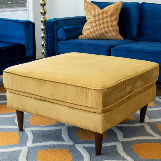 The Amber Square Upholstered Ottoman Linen with wooden legs sits on a patterned rug in front of a blue sofa with a yellow pillow, complemented by a gold floor lamp nearby.