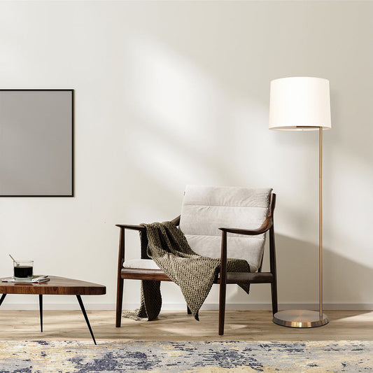A modern living room includes a cushioned wooden chair with a green throw, a Zenith Offset Base Floor Lamp with a drum-shaped linen shade in gold/white, a rectangular coffee table holding a drink and book, and a blank framed picture on the wall.