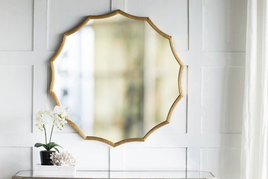 A Round Sunburst Wall Mirror with a gold frame hangs on a white paneled wall above a table holding a potted orchid, decorative coral, and stacked books. Light enters from the right, highlighting this stylish entryway decor.