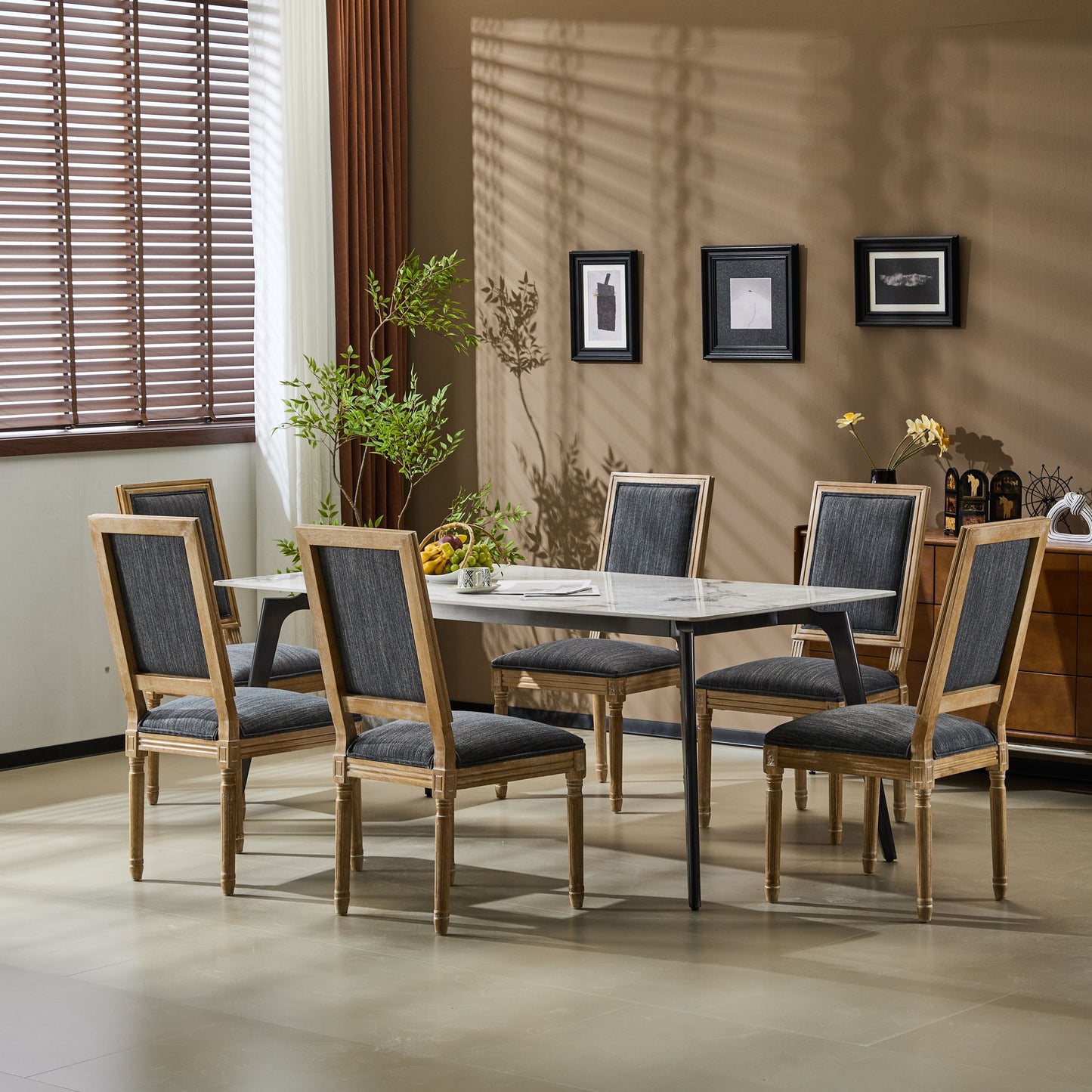 A modern dining room features a rectangular table, six French Country Style Dining Chairs with upholstered seating and fluted legs, a sideboard, potted plants, fruit on the table, framed art on a beige wall, and sunlight streaming through window blinds.