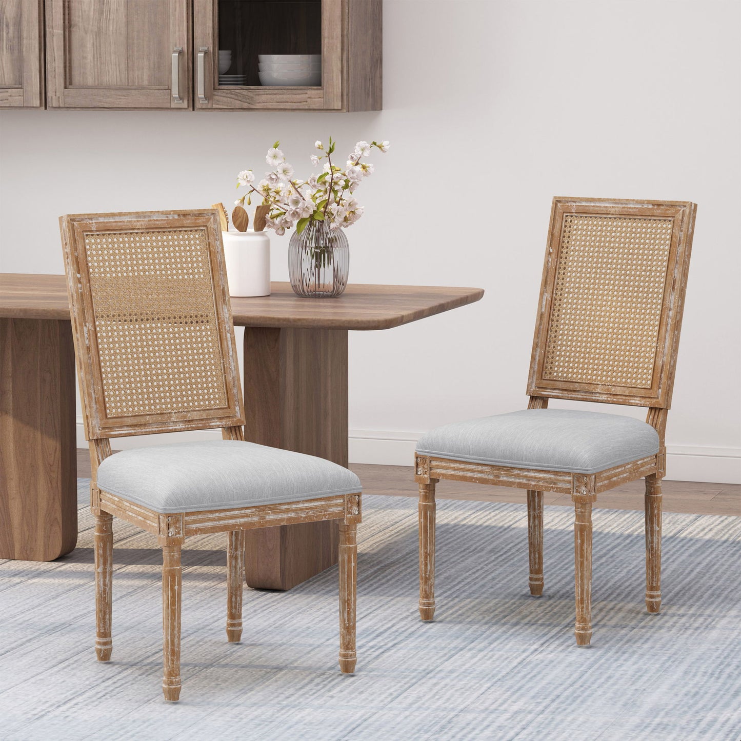 Two French Country Dining Chairs with rectangular cane backrests and smooth upholstered seats are set on a light blue rug in the dining area, near a wooden table with a vase of flowers and a cabinet in the background.