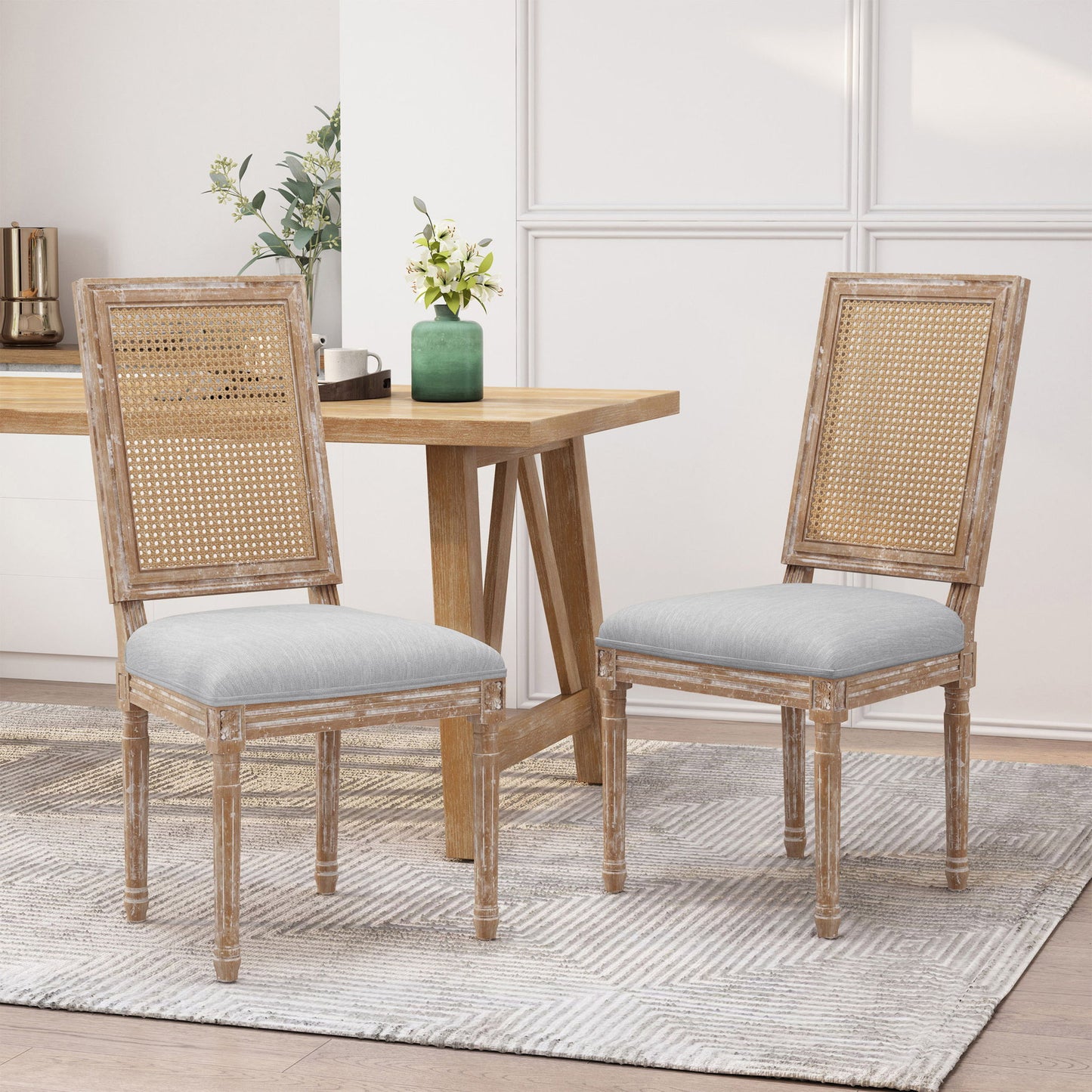 Two French Country Dining Chairs with rectangular cane backrests and smooth upholstered seats sit on a light patterned rug by a wooden dining table. A green vase with flowers is on the table, set against white paneled walls.