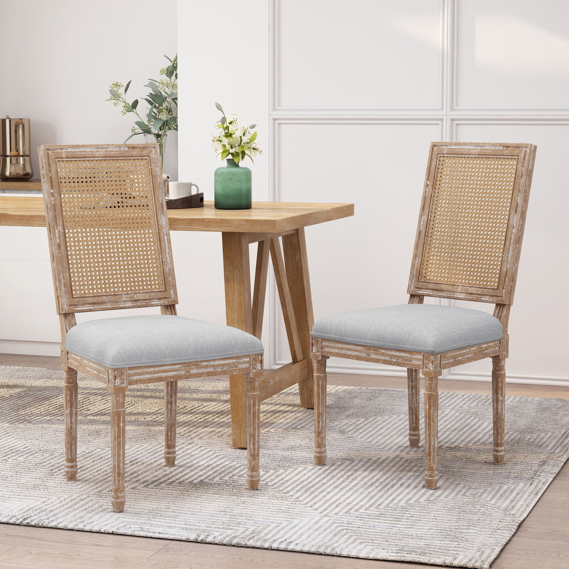 Two French Country Dining Chairs with rectangular cane backrests and smooth upholstered seats sit on a light patterned rug by a wooden dining table. A green vase with flowers is on the table, set against white paneled walls.