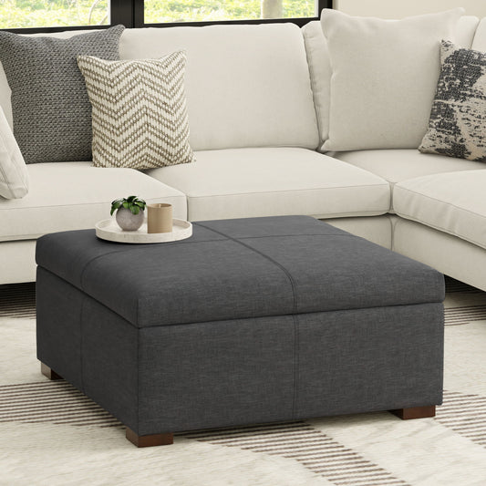 A modern living room features the Gabbie Coffee Table Upholstered Storage Ottoman in dark gray on a light rug, topped with a tray, plant, and decor. Behind it sits a light sectional sofa adorned with patterned and solid pillows.