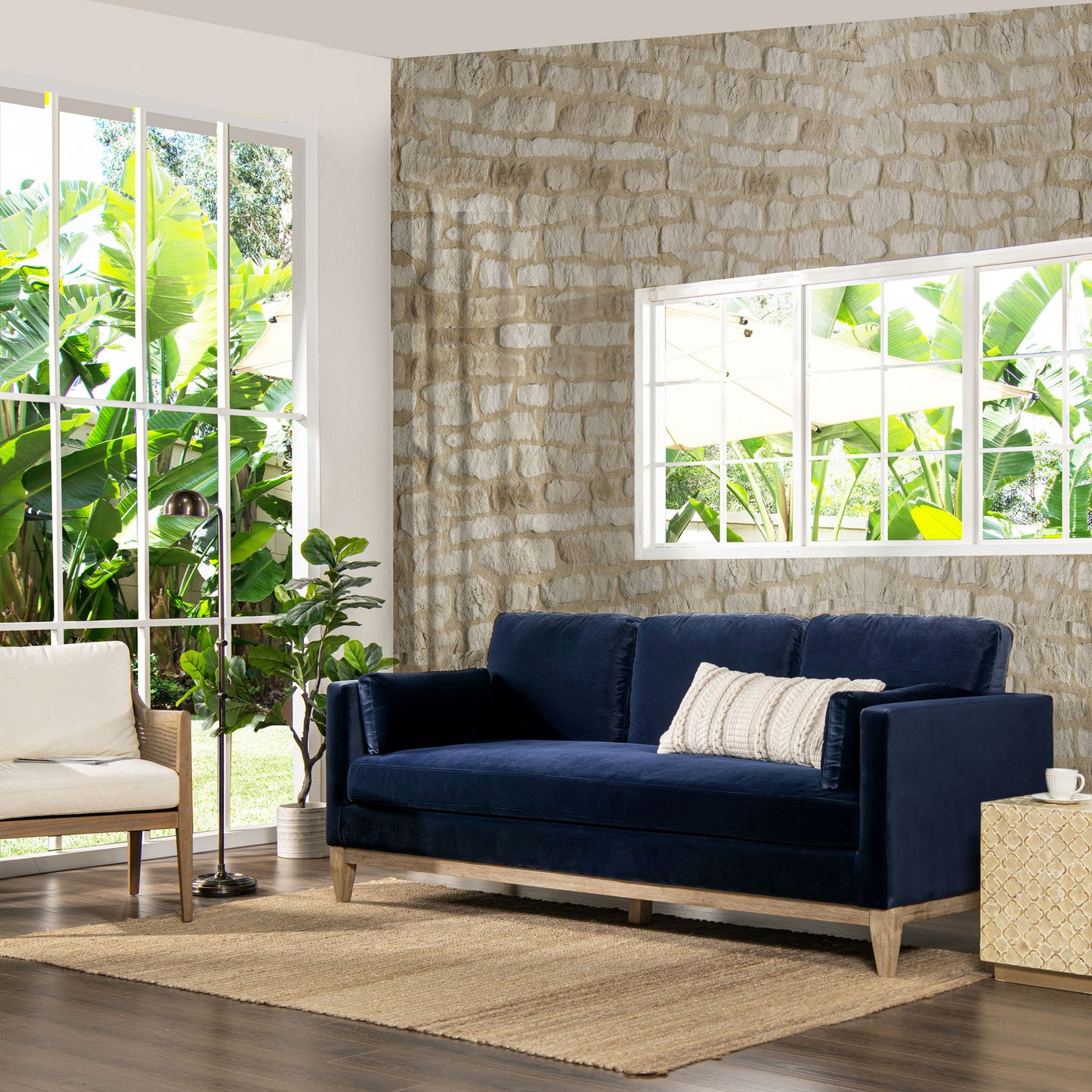 A modern living room featuring the Knox - Modern Farmhouse Sofa in navy blue, a white armchair, woven rug, indoor plants, and large windows framing lush greenery. One wall is adorned with light-colored stone.