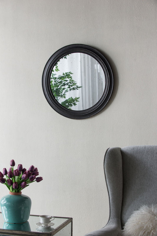 A Circle Wall Mirror With Wooden Frame, Antique Classic Accent Mirror in black hangs on a light wall, reflecting trees. Below, a glass table with purple tulips and a teacup sits beside a gray armchair with a fuzzy white pillow.