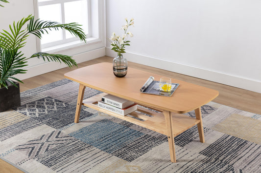The Bahamas - Coffee Table, styled with books, a white flower vase, a rolled magazine, and a glass of juice, stands on a patterned rug in a bright modern living room with a large potted plant near the window.