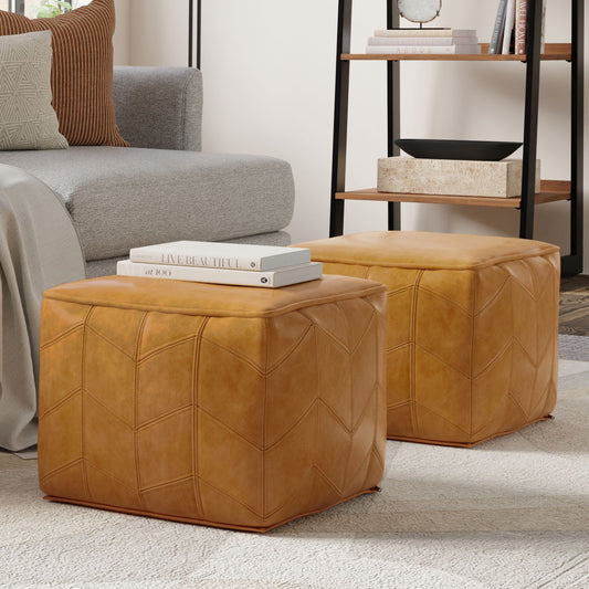 Two Morden Square Pouf ottomans in brown sit on a light rug in a modern living room, with a grey sofa, stacked books, and a black shelving unit in the background.