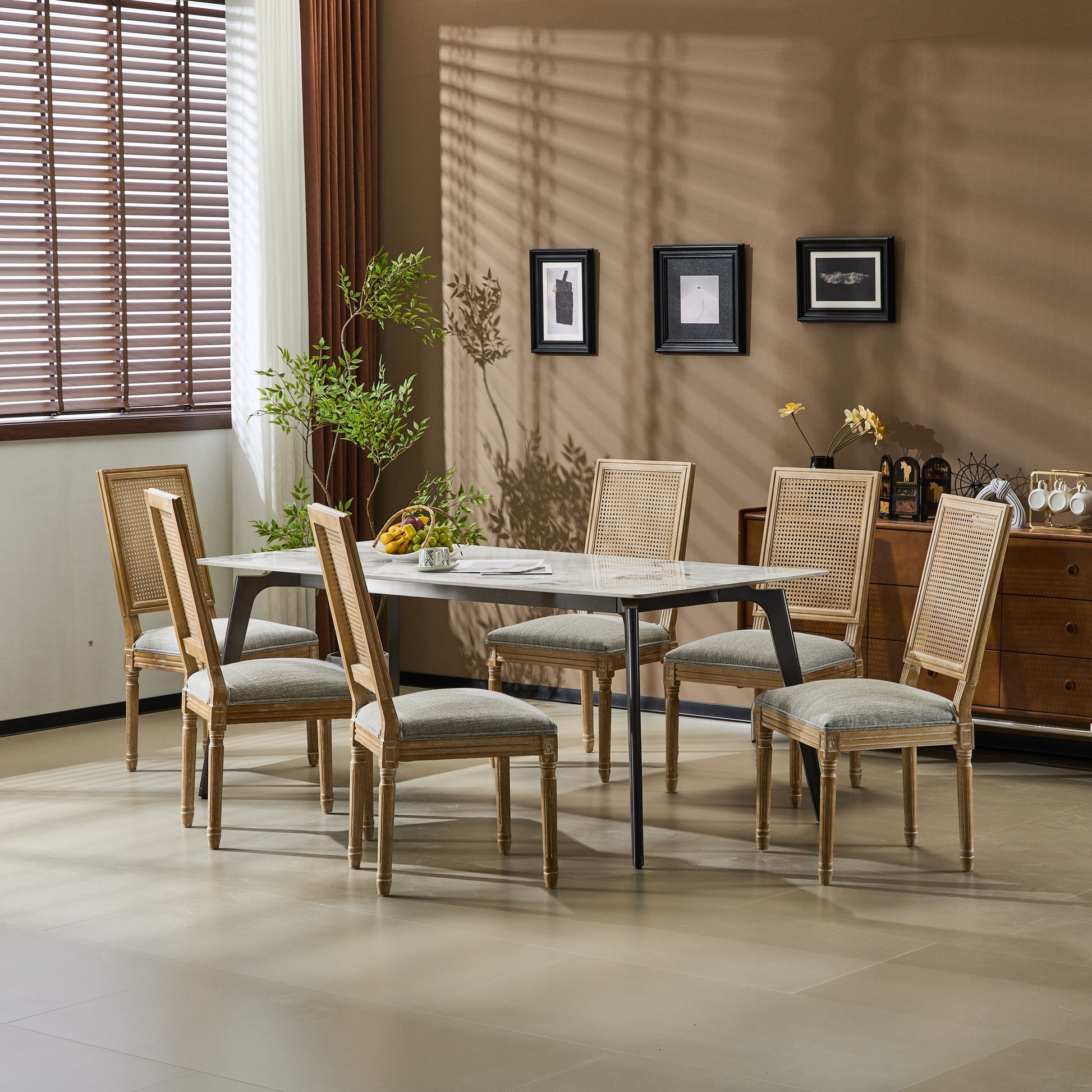 A modern dining room features a rectangular white table with six French Country Dining Chairs, each with a rectangular cane backrest and smooth upholstered seating, set by a sideboard, wall art, plants, and sunlight through window blinds.