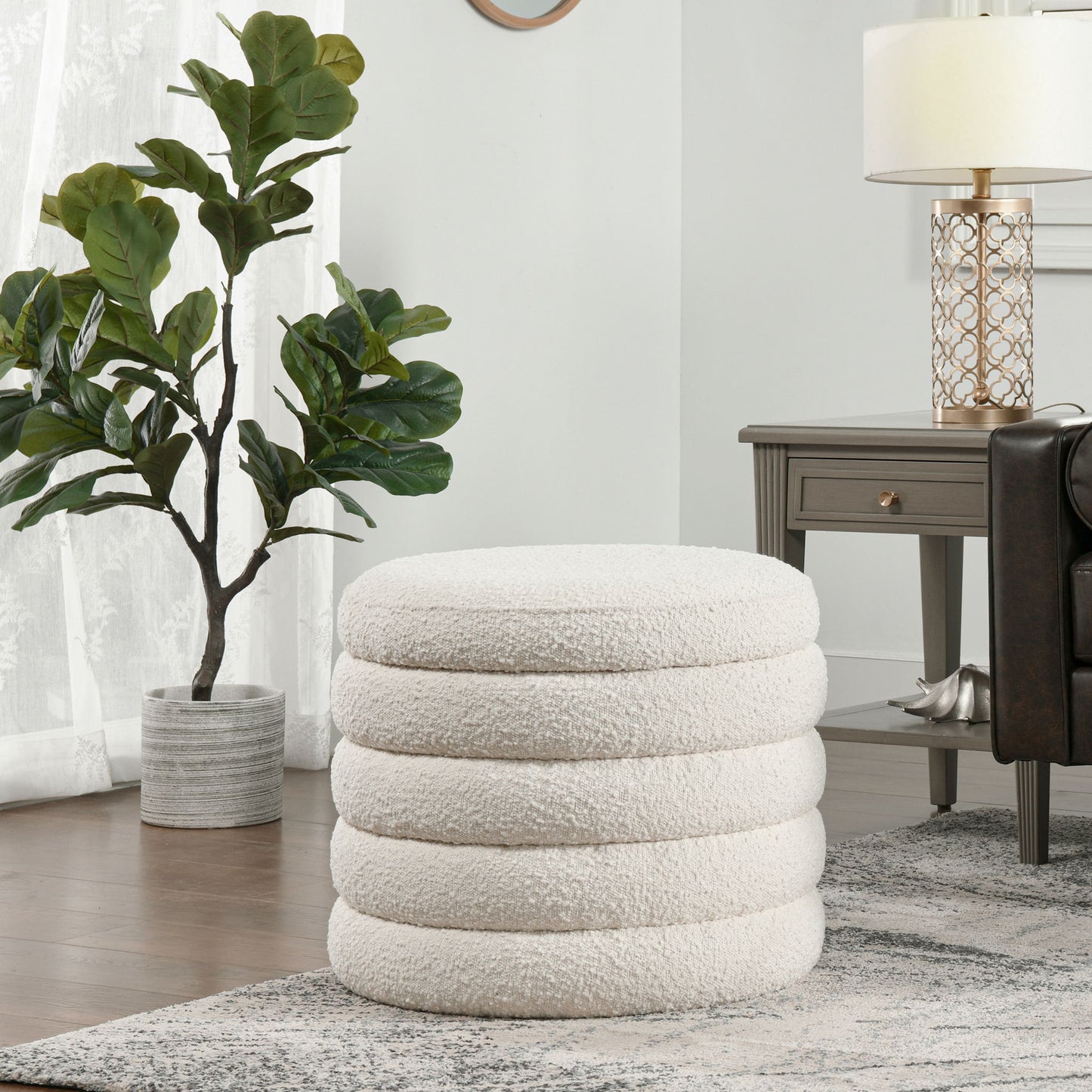The Fuji Oversized Round Storage Ottoman, featuring a plush white design with stacked rings, sits on a patterned rug in a modern living room beside a dark wood side table and a potted plant near sheer-curtained windows.