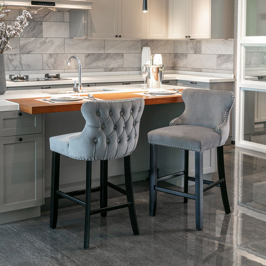 Two Contemporary Velvet Upholstered Wing-Back Barstools with button tufting and chrome nailhead trim sit at a modern kitchen island with a wooden countertop, surrounded by white cabinetry and a gray marble tile backsplash.