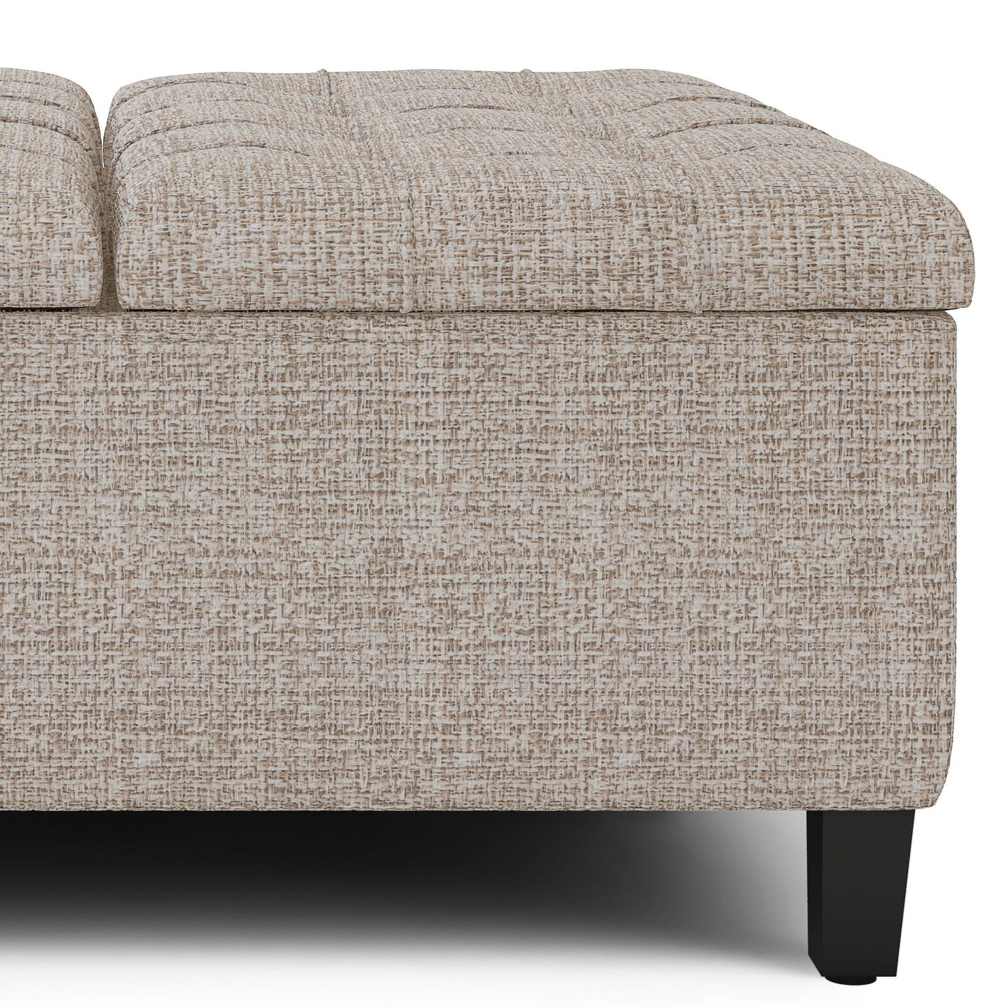 A close-up of the Harrison Upholstered Coffee Table Storage Ottoman features textured beige fabric, a tufted rectangular top, and black wooden legs, all shown against a white background.