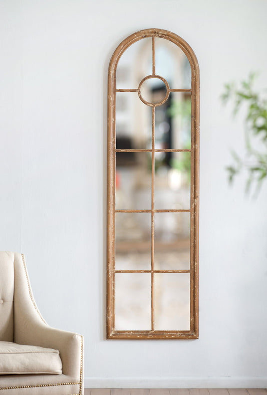 The Half Round Elongated Mirror With Decorative Window Look features a classic arched design and solid fir wood frame in brown, mounted on a white wall beside a beige chair, with a blurred reflection and green plant in the background.