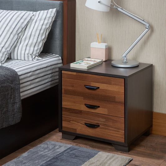 The Eloy - Accent Table in Walnut / Espresso features three wooden drawers and is styled with a silver desk lamp, reed diffuser, and closed book. It stands beside a bed with striped sheets and a gray blanket.