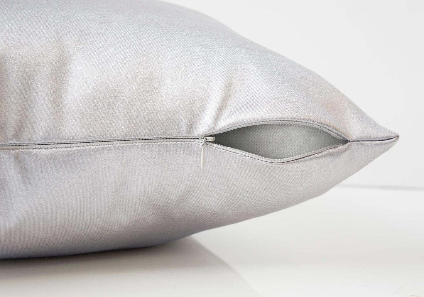 A close-up of a modern square decorative throw pillow with hypoallergenic insert, silver satin cover, and partially open visible zipper revealing the inner lining, displayed on a smooth white surface against a plain background.