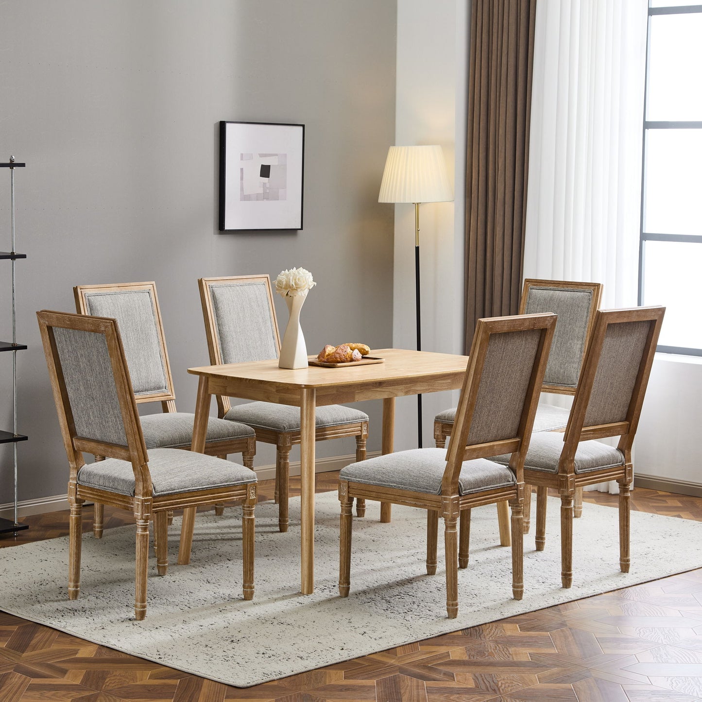 A modern dining room features a wooden table, six French Country Style Dining Chairs with upholstered seating and fluted legs, a floor lamp, framed abstract art on a gray wall, large curtained window, plus a vase and pastries on the table over a light rug.