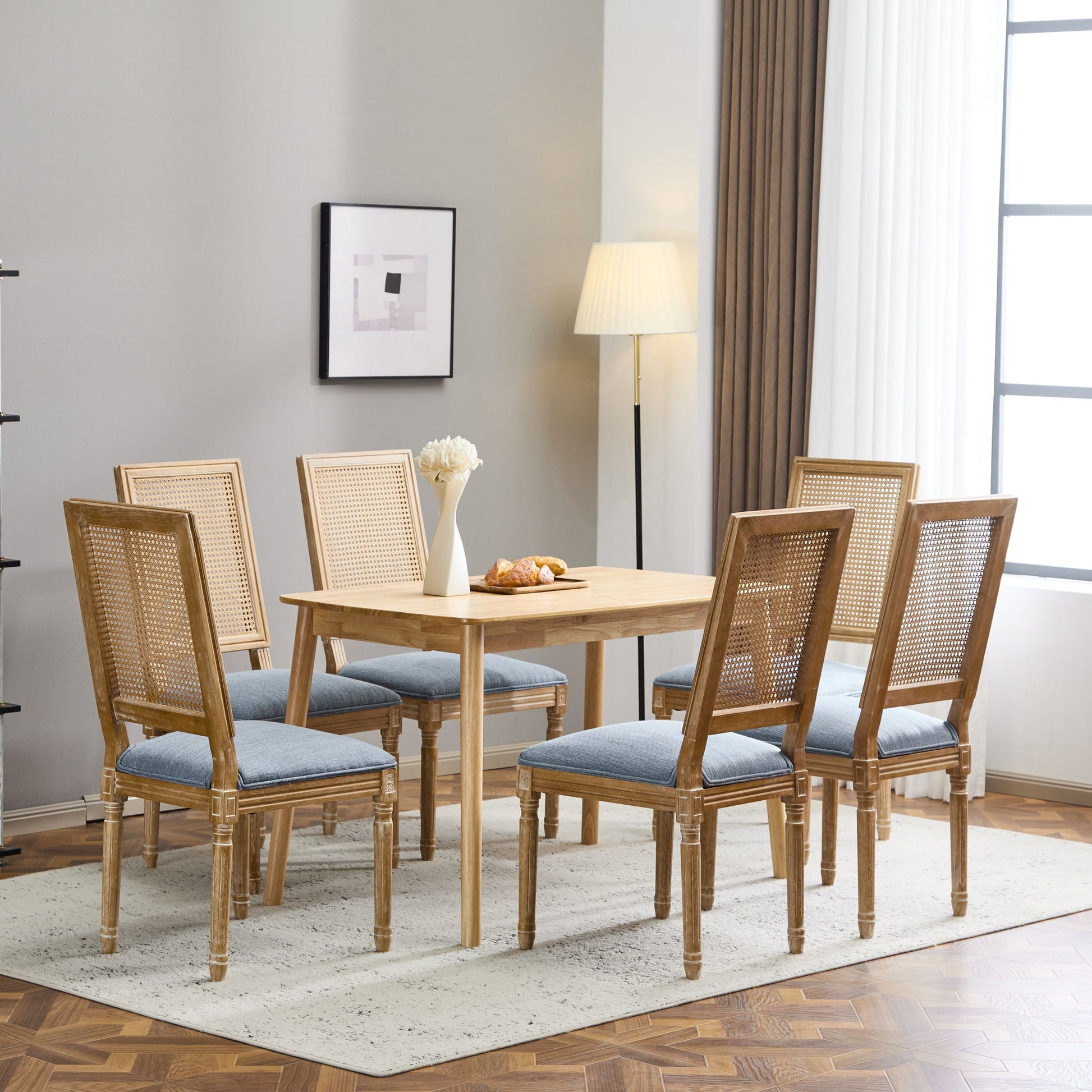 A dining room with a wooden table, six French Country Dining Chairs with rectangular cane backrests and blue upholstered seats, a floor lamp, framed wall art, white curtains on the window, and a vase of flowers plus pastries on the table.