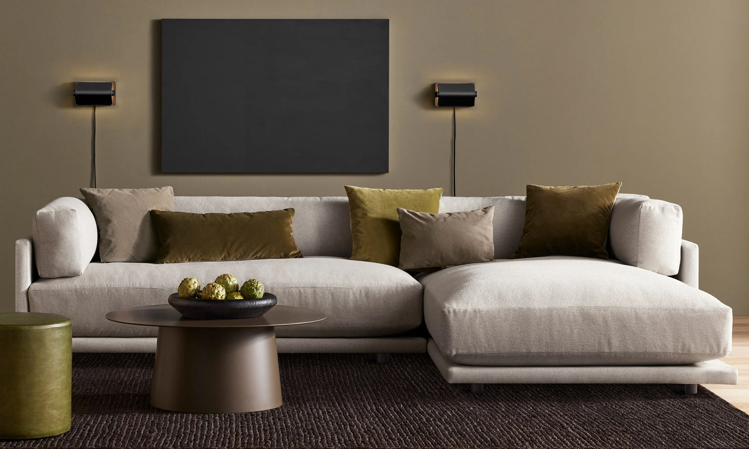 Modern living room with a beige sectional sofa adorned with green, gray, and beige pillows. A round coffee table holds a decorative centerpiece, and a black wall-mounted panel with two sconces is above the sofa. The room features earthy tones and minimalist decor.