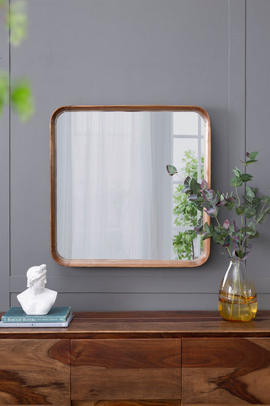 A Square Mirror With Wood Frame, Wall Mirror For Living Room Bathroom Entryway - Brown hangs on a gray wall above a wooden cabinet, with books, a white bust, and a glass vase holding green and purple foliage atop the cabinet.