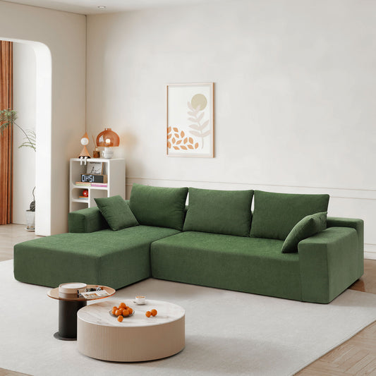 A light-filled living room features a green Modular Sectional Sofa Set in modern minimalist style, two cushions, a round coffee table with oranges, a white decor shelf, and abstract wall art.