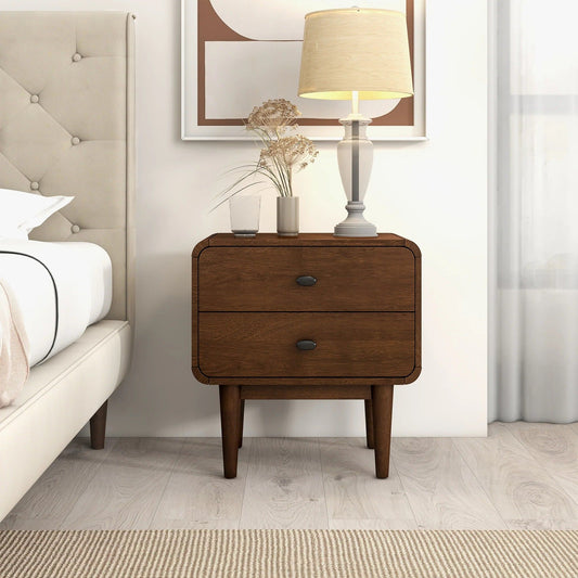 The Alexa Mid-Century Modern Style Night Stand in brown stands by the bed, topped with a beige lamp, glass vase of dried flowers, and a small glass. Minimalist artwork is above, and a window sits to the right.
