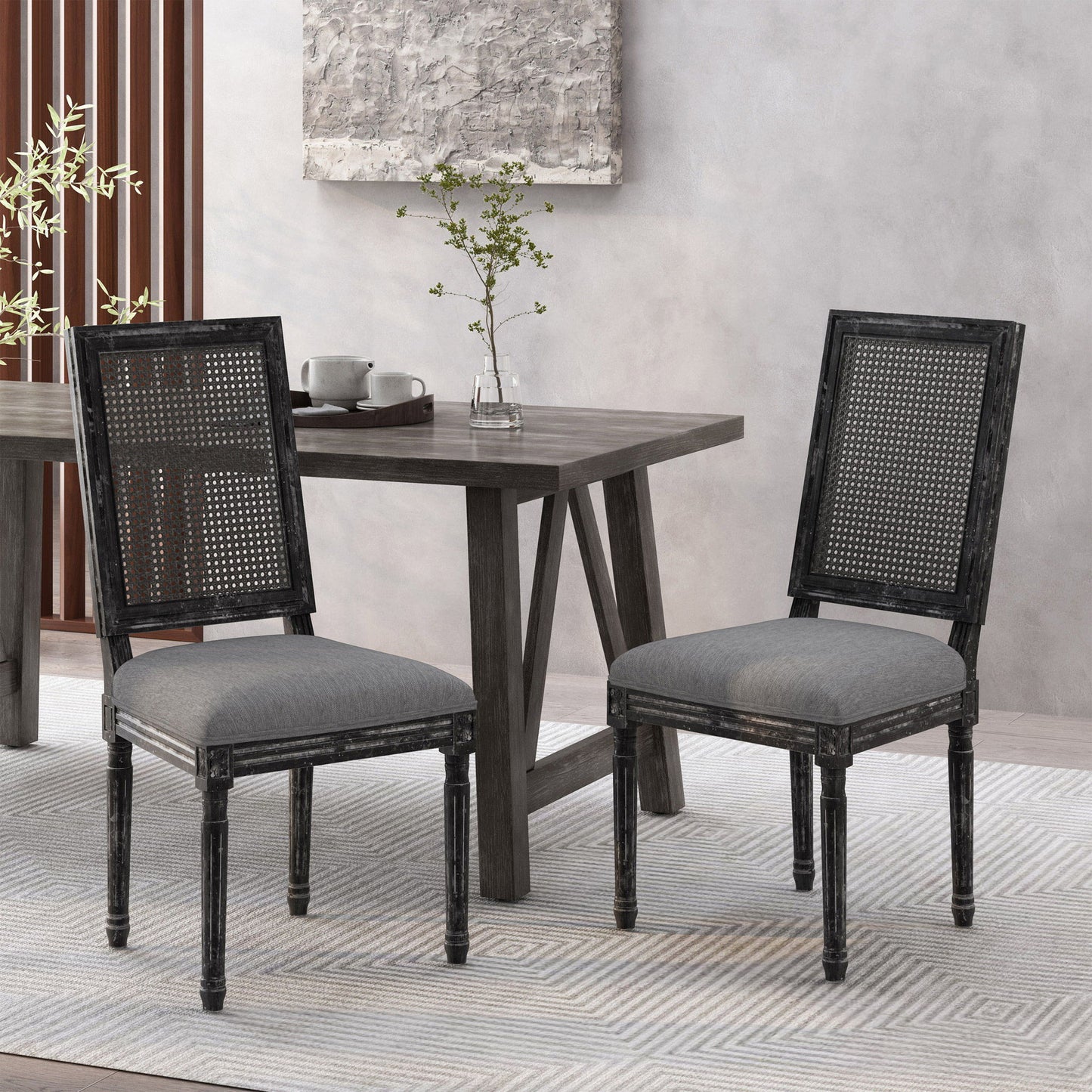 Two French Country Dining Chairs with rectangular cane backrests and smooth upholstered seats sit in front of a modern wooden dining table topped with a vase of greenery and tea set, against a neutral wall and rug.