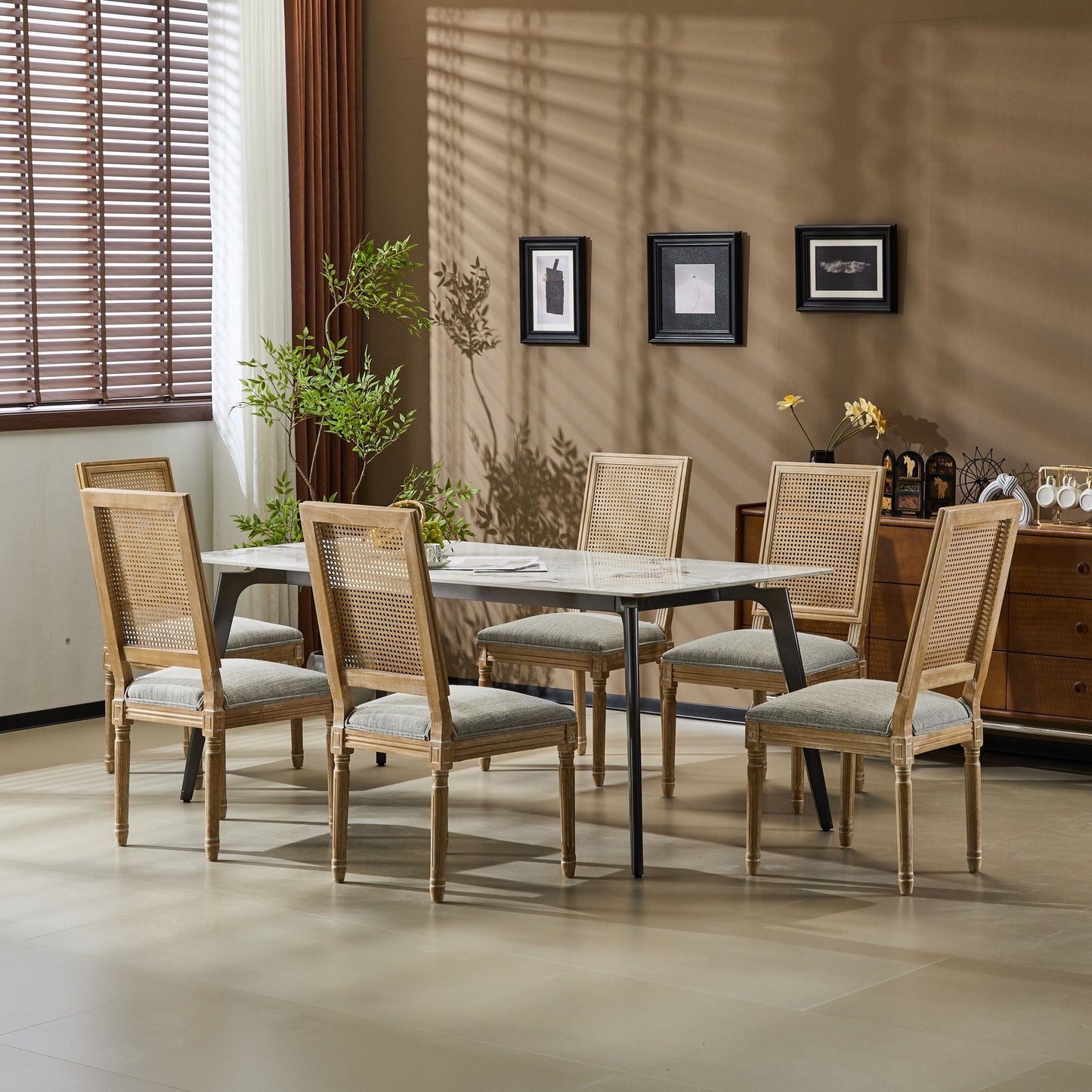 A modern dining room features a rectangular table, six French Country Dining Chairs with cane backrests and upholstered seats, a corner plant, wall art, and a sideboard set against a tan wall lit by sunlight through window blinds.
