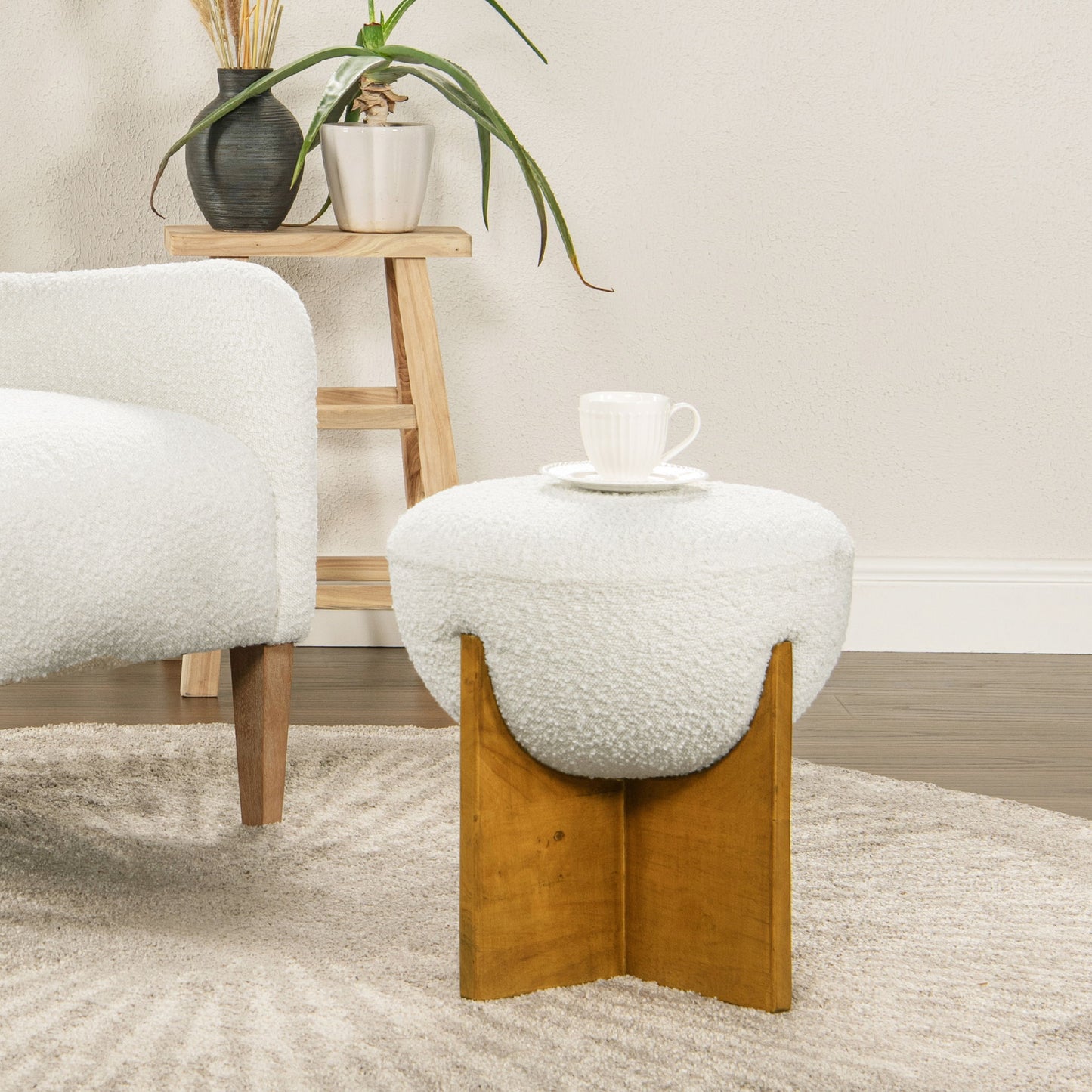 A modern living room showcases the Bali - Round Upholstered Ottoman with a wooden base, used as a side table for a white teacup and saucer, placed next to a white sofa and a wooden shelf with plants.