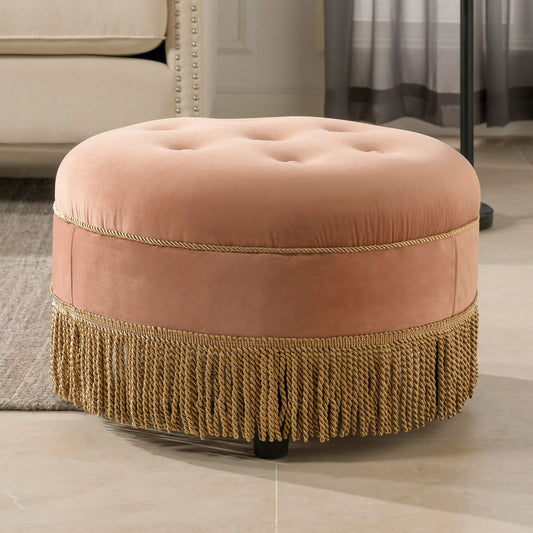 The Yolanda - Round Upholstered Accent Ottoman, featuring pink velvet fabric, gold braided trim, and fringe at the base, sits on a tile floor in a neutral-toned living room.