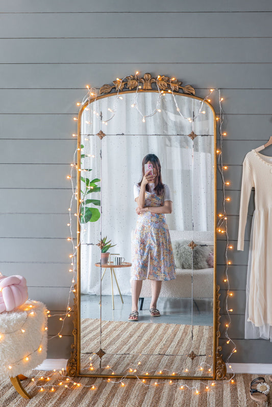 A person takes a selfie in front of the Lavish Full Length Mirror, an antique gold metal floor mirror. The room features gray paneled walls, a soft chair, a hanging dress, a plant, and cozy decor with string lights on the mirror.