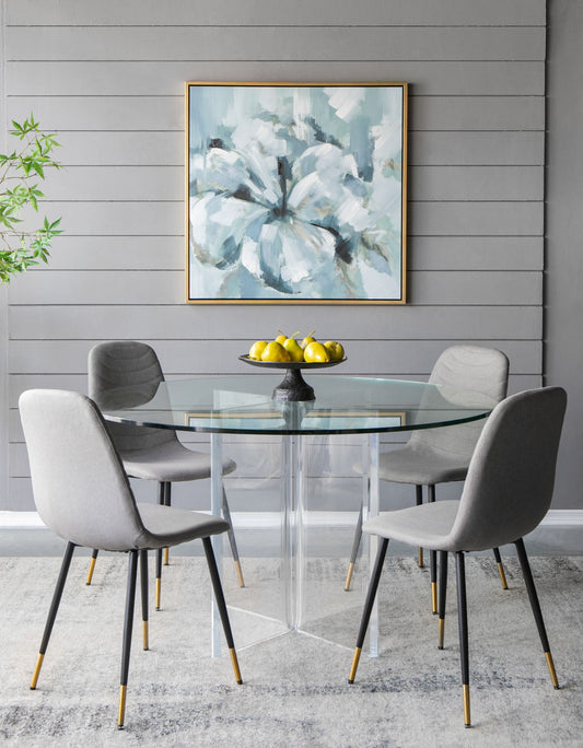 A modern dining area featuring a round glass table, four gray chairs with gold-tipped legs, a bowl of yellow lemons, a Large Modern Flower Oil Painting, SquareFrame Wall Art - Blue / Gray on the gray paneled wall, and a plant in the corner.