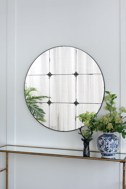 A Traditional Decor Style Round Antique Glass Wall Mirror hangs above a gold console table holding a blue-and-white floral vase with green leafy branches, a black deer figurine, and another plant reflected in the mirror.