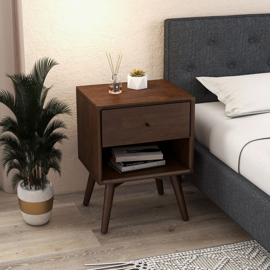 The Caroline Mid-Century Modern Style Night Stand in brown, featuring angled legs, stands beside a gray upholstered bed. On it are a diffuser, candle, and small plant; books are stored below. A potted plant sits nearby on the floor.
