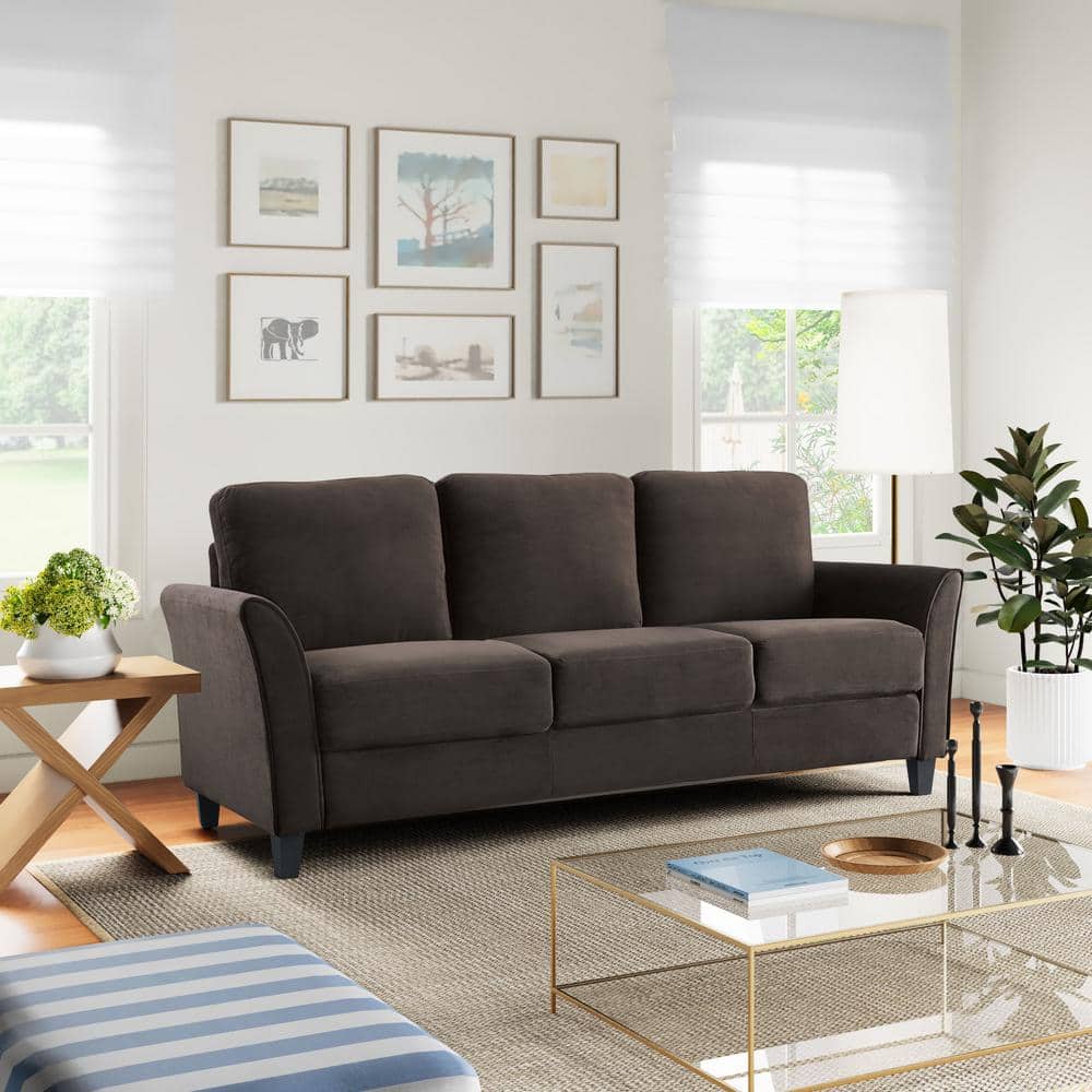 A modern living room features the Lifestyle Solutions Wesley 80.3 in. dark grey sofa, a wooden side table with plants, glass coffee table with books, striped ottoman, floor lamp, framed wall art, and large windows bringing in natural light.