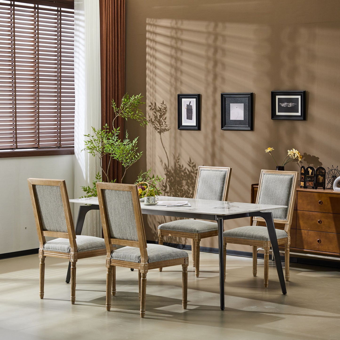 A modern dining room featuring a rectangular marble table, five French Country Style Dining Chairs with upholstered seating and fluted legs, a potted plant, wall art, a sideboard, and sunlight casting shadows through wooden blinds.