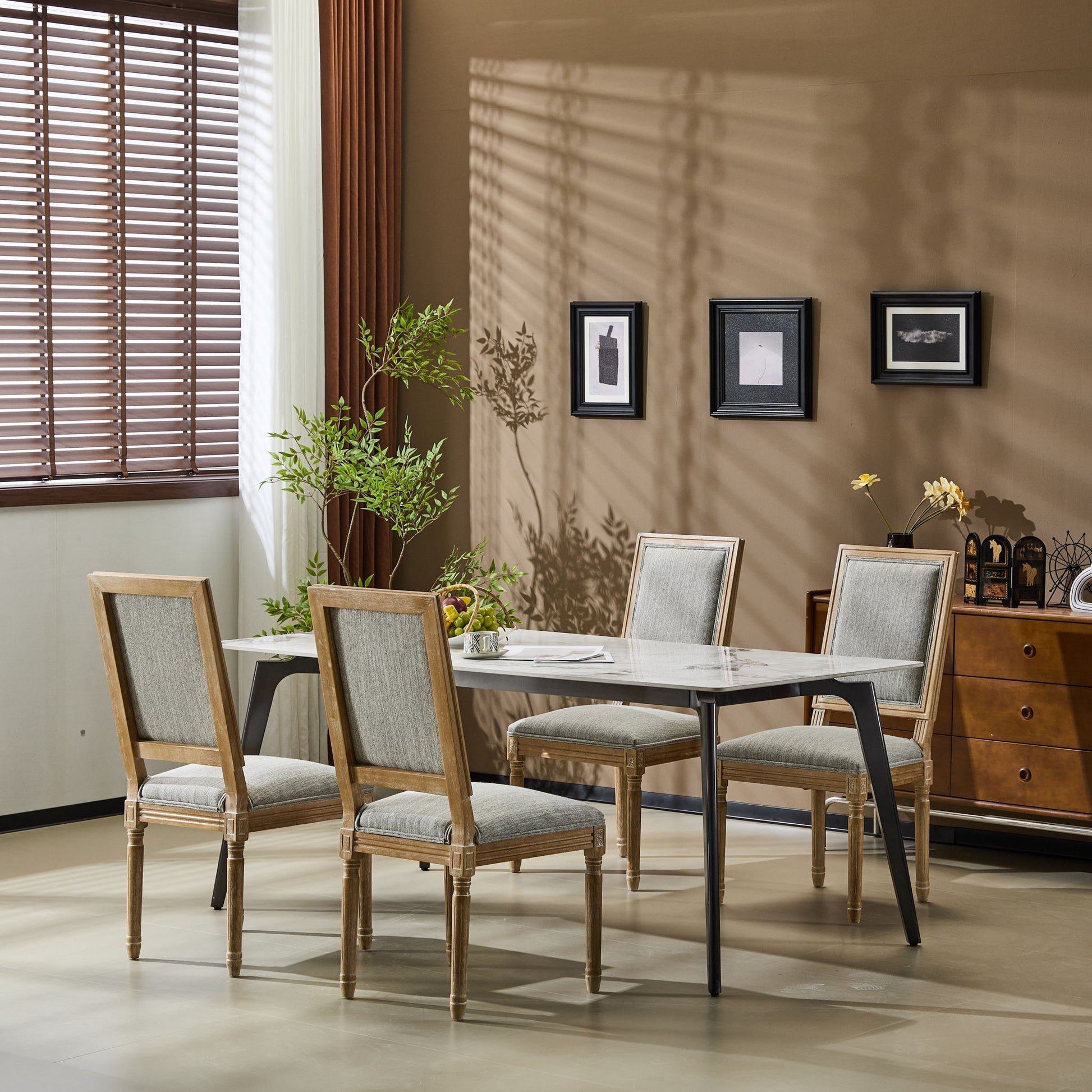 A modern dining room featuring a rectangular marble table, five French Country Style Dining Chairs with upholstered seating and fluted legs, a potted plant, wall art, a sideboard, and sunlight casting shadows through wooden blinds.