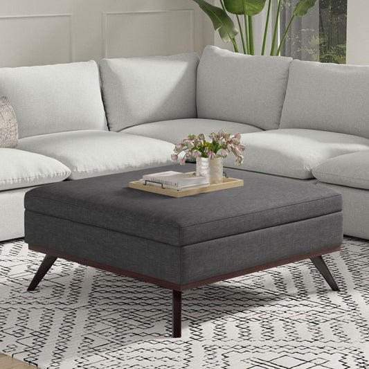 The Owen Multifunctional Square Coffee Table Storage Ottoman with wooden legs sits on a patterned rug in front of a light gray sectional, topped with a tray, books, and pink flowers beside a green potted plant in a modern living room.
