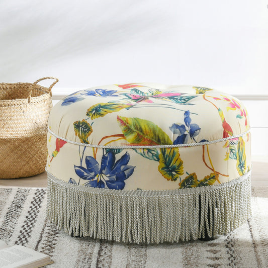 The Yolanda Round Upholstered Accent Ottoman, featuring a colorful floral print and silver fringed trim, sits on a striped rug beside a woven basket in a bright room.