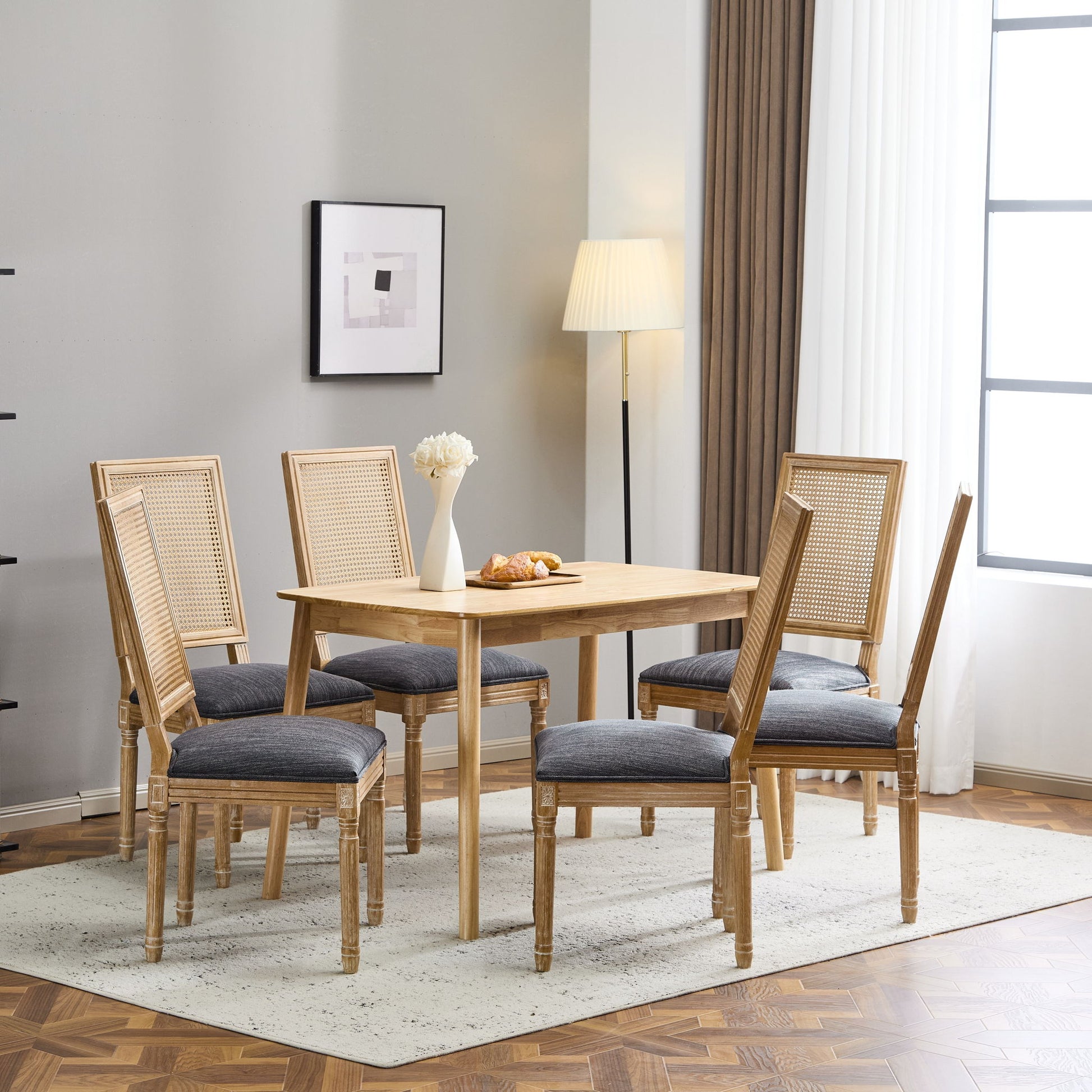 A modern dining room with a wooden table, six French Country Dining Chairs featuring rectangular cane backrests and smooth upholstered seats, a floor lamp, framed wall art, and a vase of white flowers on the table atop a light rug.