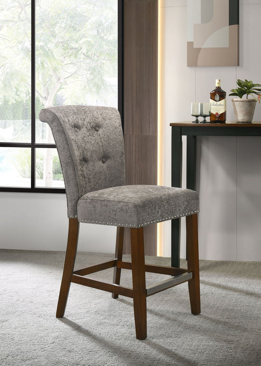 The Auggie 20.5" Fabric Counter Height Chair with nailhead trim features gray upholstery and wooden legs, positioned on a light carpet near a window and a table holding bottles, glasses, and a potted plant.
