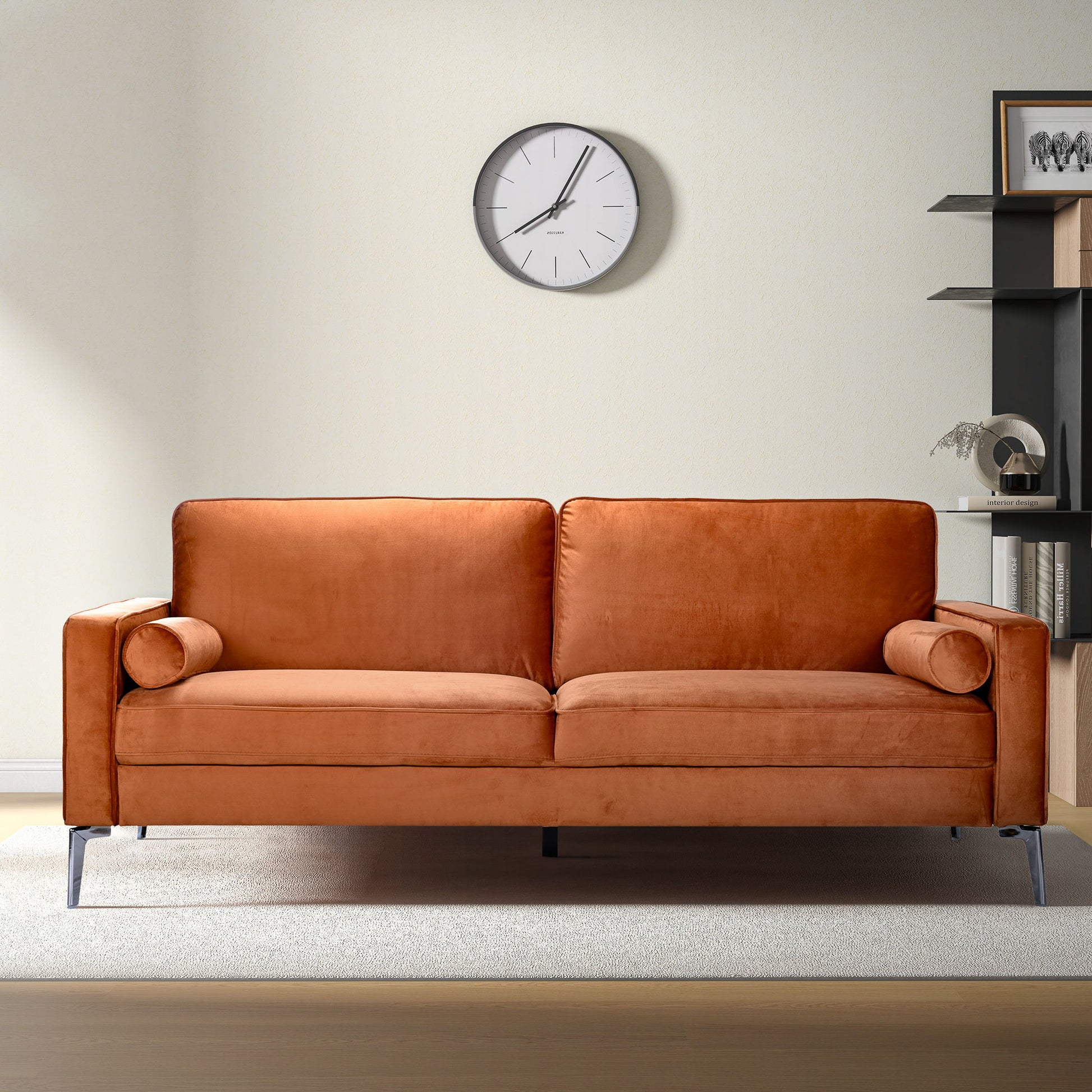 A modern living room featuring the Axten - Sofa - Orange with two matching bolster pillows, a light rug, a wall clock, and black shelves with decor against a cream-colored wall.