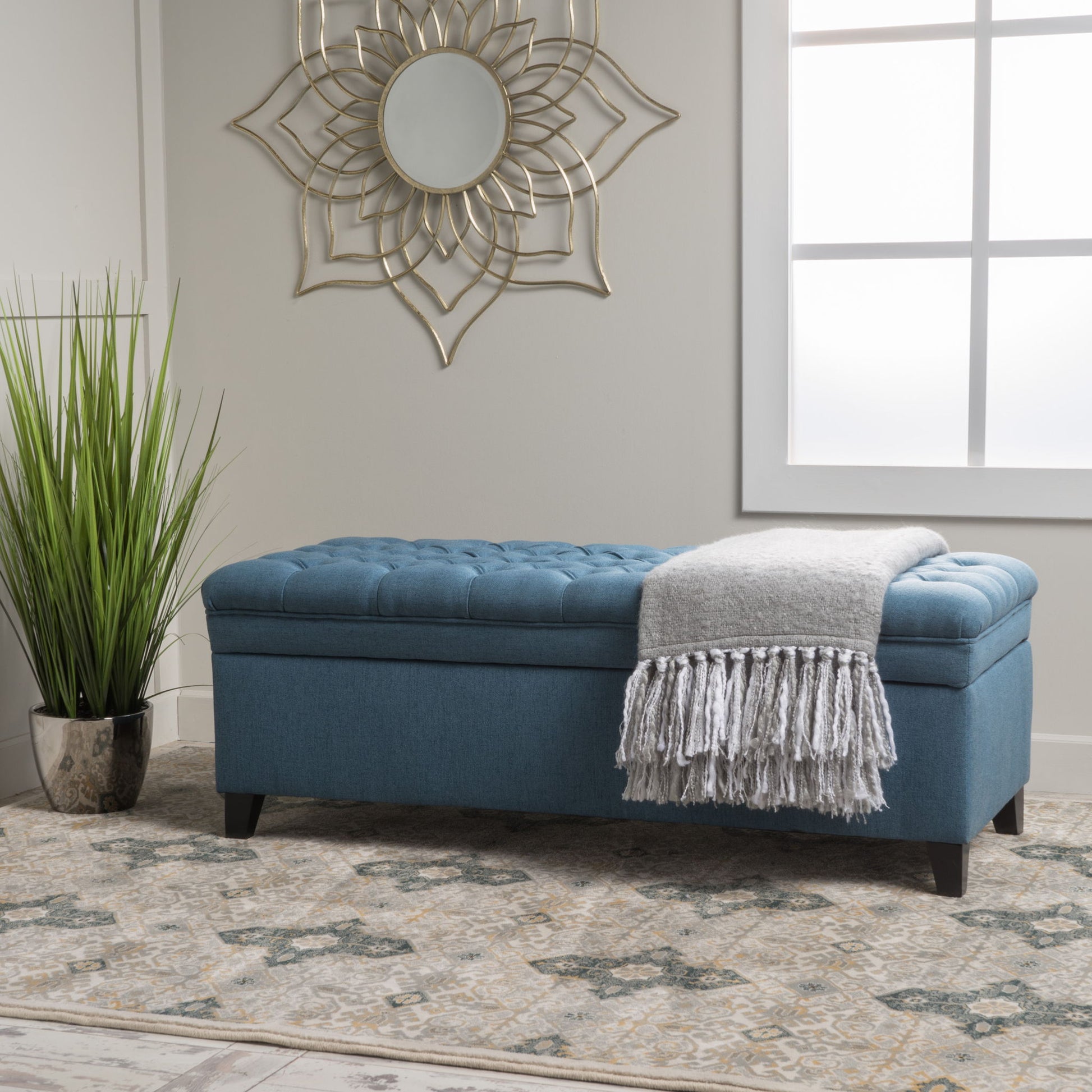 The Juliana Storage Ottoman with Birch Legs sits on a patterned rug near a window, topped with a gray fringed throw blanket. Beside it stands a tall potted plant, while a decorative gold mirror hangs on the wall above.
