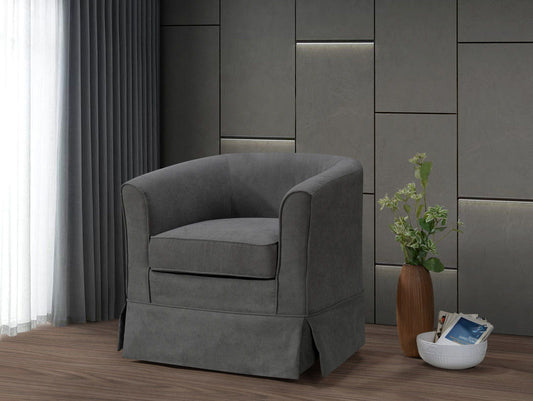 The Tucker Woven Fabric Swivel Barrel Chair in gray sits on a wooden floor by a wall with geometric panels, accompanied by a tall vase of green flowers and a white magazine bowl. Sheer curtains gently filter natural light into the space.