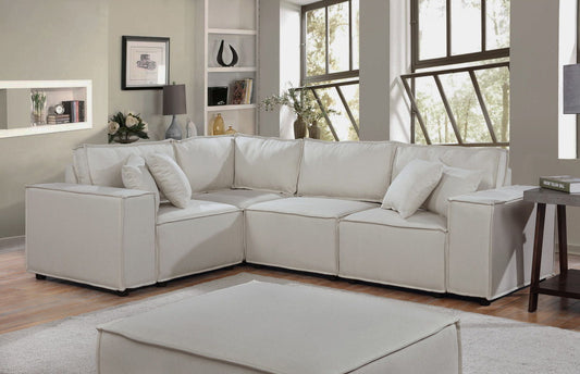 A modern living room featuring the Melrose Modular Sectional Sofa with Ottoman in white. Large windows, light walls, bookshelves, a table lamp, and decor complement the hardwood floors and white rug.