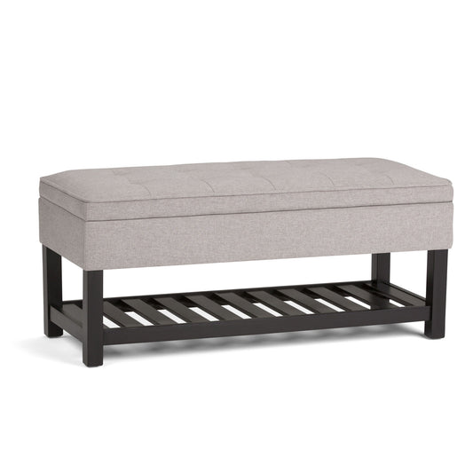 The Cosmopolitan Storage Ottoman Bench features a light gray cushioned top, black wooden legs, and an open slatted shelf underneath for added storage.