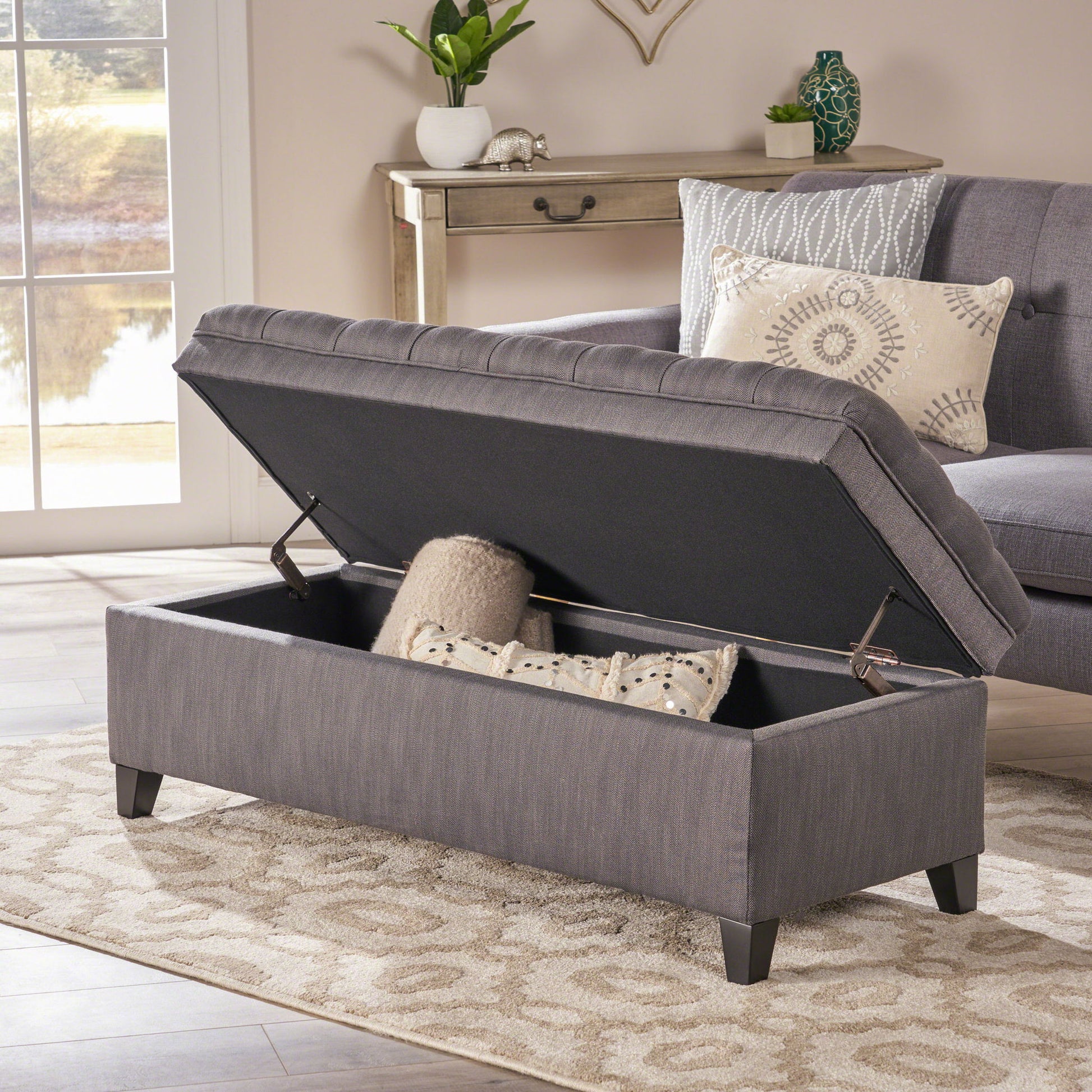 The Juliana Storage Ottoman With Birch Legs, shown open to reveal two pillows inside, sits on a beige patterned rug in a living room beside a gray sofa and window with an outdoor view.