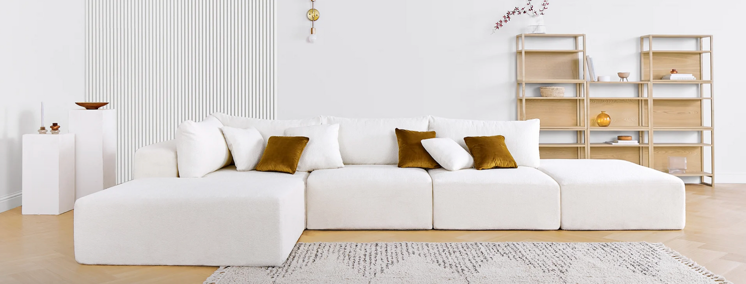 A minimalist living room featuring a large white sectional sofa adorned with white and mustard yellow throw pillows. Behind the sofa is a wooden shelving unit with decorative items. The room has light wooden flooring and a patterned rug.