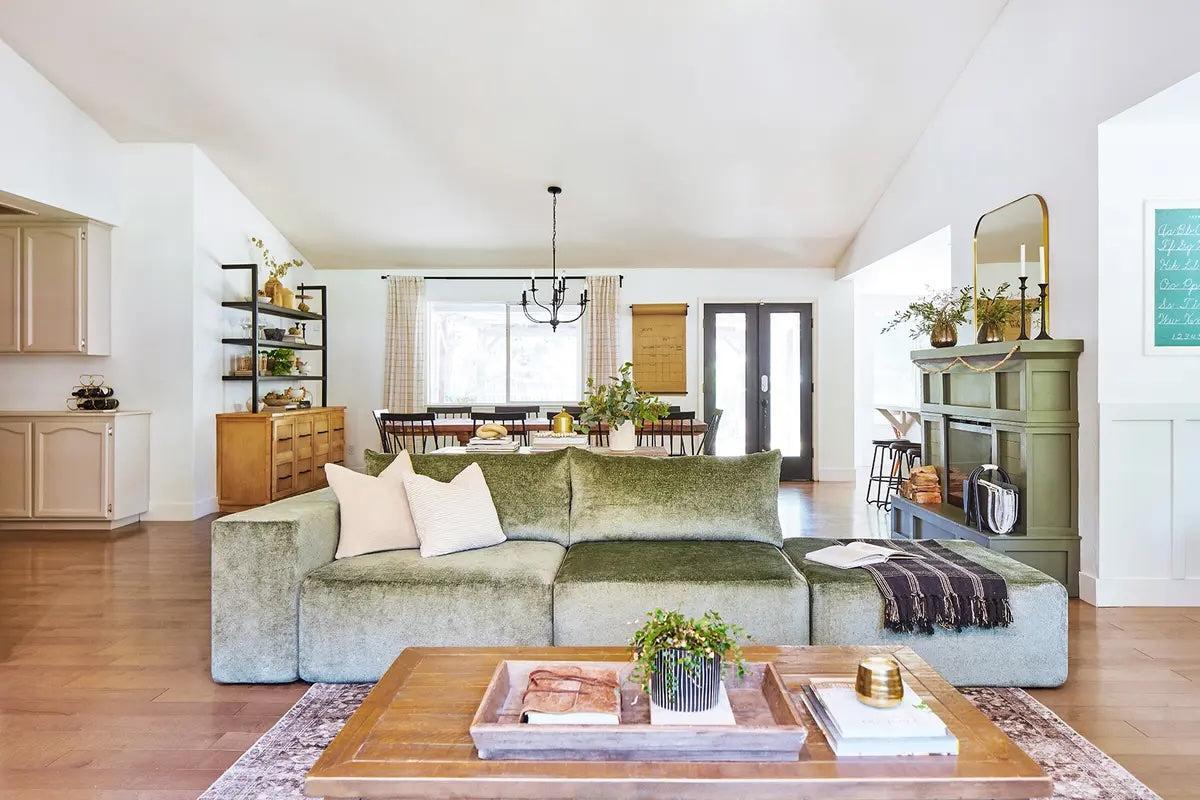 A stylish open-concept living and dining area with a green sectional sofa, wooden coffee table, and light-colored furnishings. The room features large windows, modern light fixtures, and a mix of traditional and contemporary décor elements.